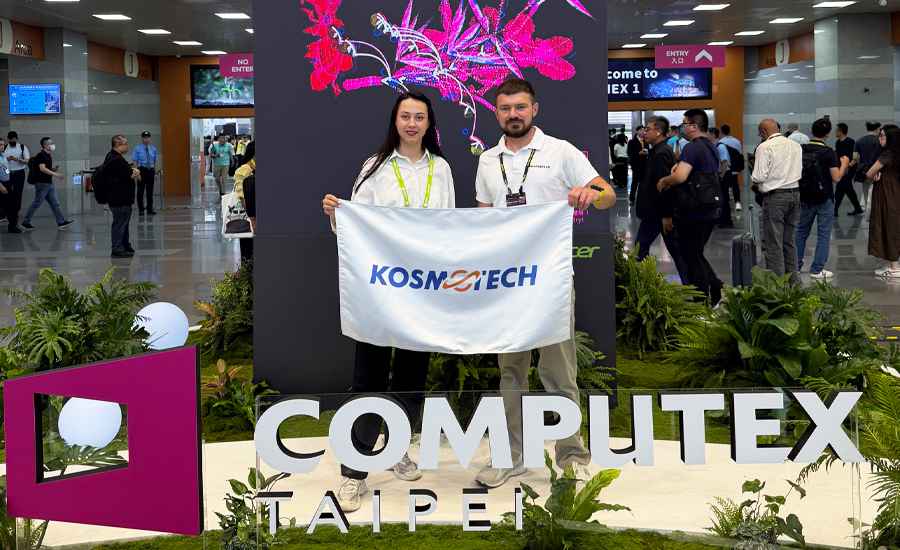 900х550рх-новина-на-сайт Представники компанії тримають прапор Kosmotech на виставці Computex Taipei. Вони стоять біля великої інсталяції з написом «COMPUTEX TAIPEI» у виставковому центрі, на фоні експозиційного стенда та відвідувачів заходу.
