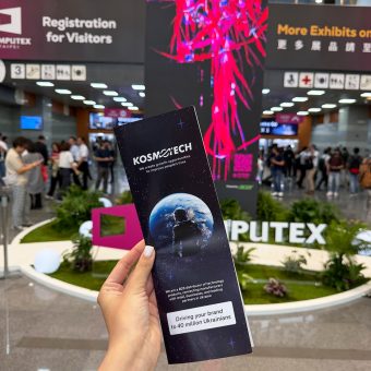 Рука тримає корпоративний буклет Kosmotech на фоні виставкового простору COMPUTEX. На задньому плані видно інсталяцію заходу, інформаційні стенди та відвідувачів міжнародної технологічної виставки.